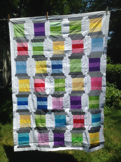 Colorful patchwork quilt resembling thread spools in red, green, blue, purple, and yellow, hanging outdoors against greenery