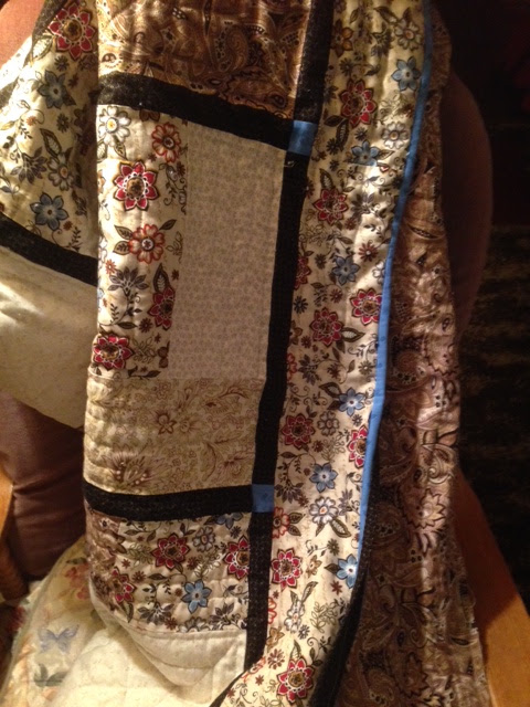 Folded quilt with floral and paisley fabric patches separated by black sashing and blue accents, draped over furniture, warm homey feel