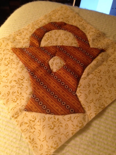 Fabric applique in the shape of a basket made from striped orange and cream patterned cloth, sewn onto a pale yellow quilt block