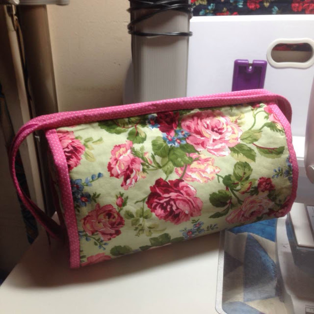 A quilted fabric bag with pink floral print and pink polka-dot trim, set on a table with household items, soft domestic mood