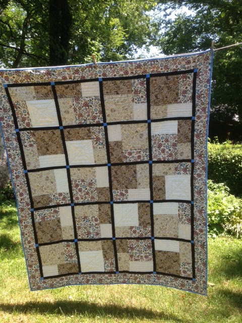 Patchwork quilt in brown, cream and floral fabric squares hung outdoors on a line against green trees and grass, warm earthy mood